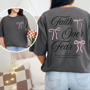 May include: Dark gray t-shirt with the text "Faith Over Fear" and pink bow accents. The back of the shirt features the text "Faith Over Fear" with three pink bows. The front of the shirt has a single pink bow. The shirt also has a Bible verse.