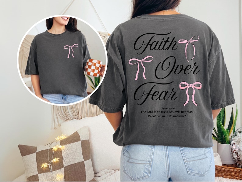 May include: Dark gray t-shirt with the text "Faith Over Fear" and pink bow accents. The back of the shirt features the text "Faith Over Fear" with three pink bows. The front of the shirt has a single pink bow. The shirt also has a Bible verse.
