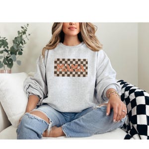 May include: A light gray sweatshirt featuring the word "MAMA" in a retro font, set against a checkered brown and tan background. The sweatshirt is a crew neck style and is made of a soft material. The overall theme is casual and comfortable.