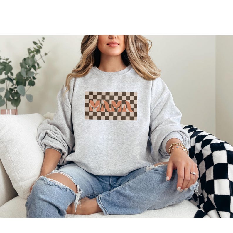 May include: A light gray sweatshirt featuring the word "MAMA" in a retro font, set against a checkered brown and tan background. The sweatshirt is a crew neck style and is made of a soft material. The overall theme is casual and comfortable.