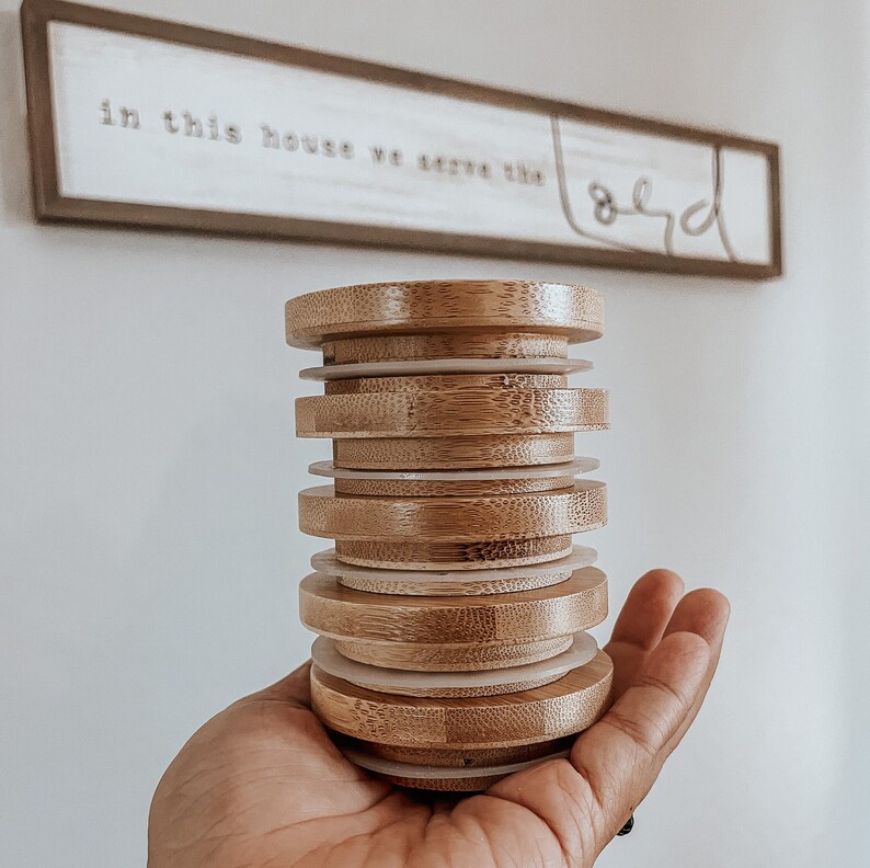 May include: A stack of eight round, wooden, bamboo lids with white rings in between each lid. The lids are stacked in a person's hand.