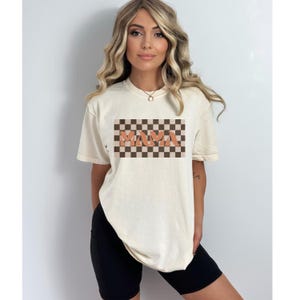May include: Cream-colored t-shirt with the word "MAMA" in orange letters on a brown and tan checkered background. The shirt has short sleeves and a relaxed fit, suitable for casual wear.