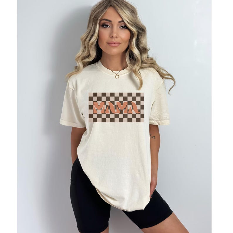 May include: Cream-colored t-shirt with the word "MAMA" in orange letters on a brown and tan checkered background. The shirt has short sleeves and a relaxed fit, suitable for casual wear.
