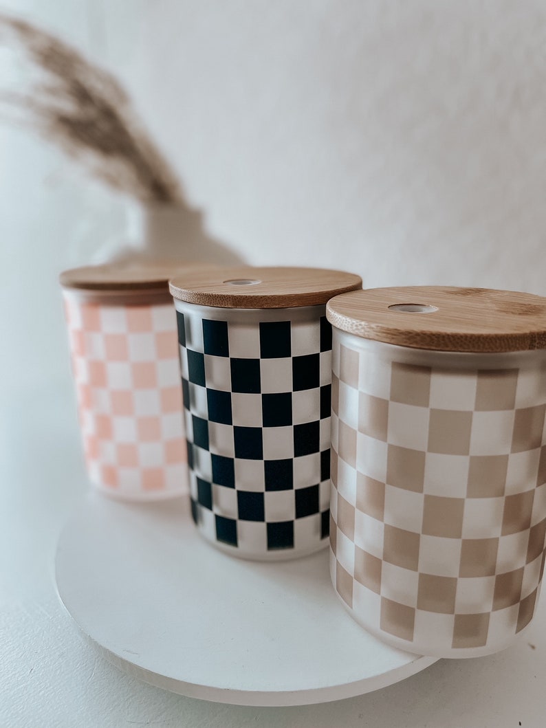 May include: Three cylindrical glass jars with wooden lids. The jars have a checkered pattern. The jars are pink, black and white, and brown and white.