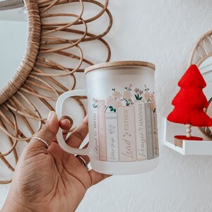 May include: A frosted glass mug with a bamboo lid and a floral design. The mug has a handle and features the words "Lead", "Worship", "Miriam", "Esther", "Prayer", "Hannah", "Legal", "Ruth", "Deborah", "Rejoice", and "Sarah" printed on the side.