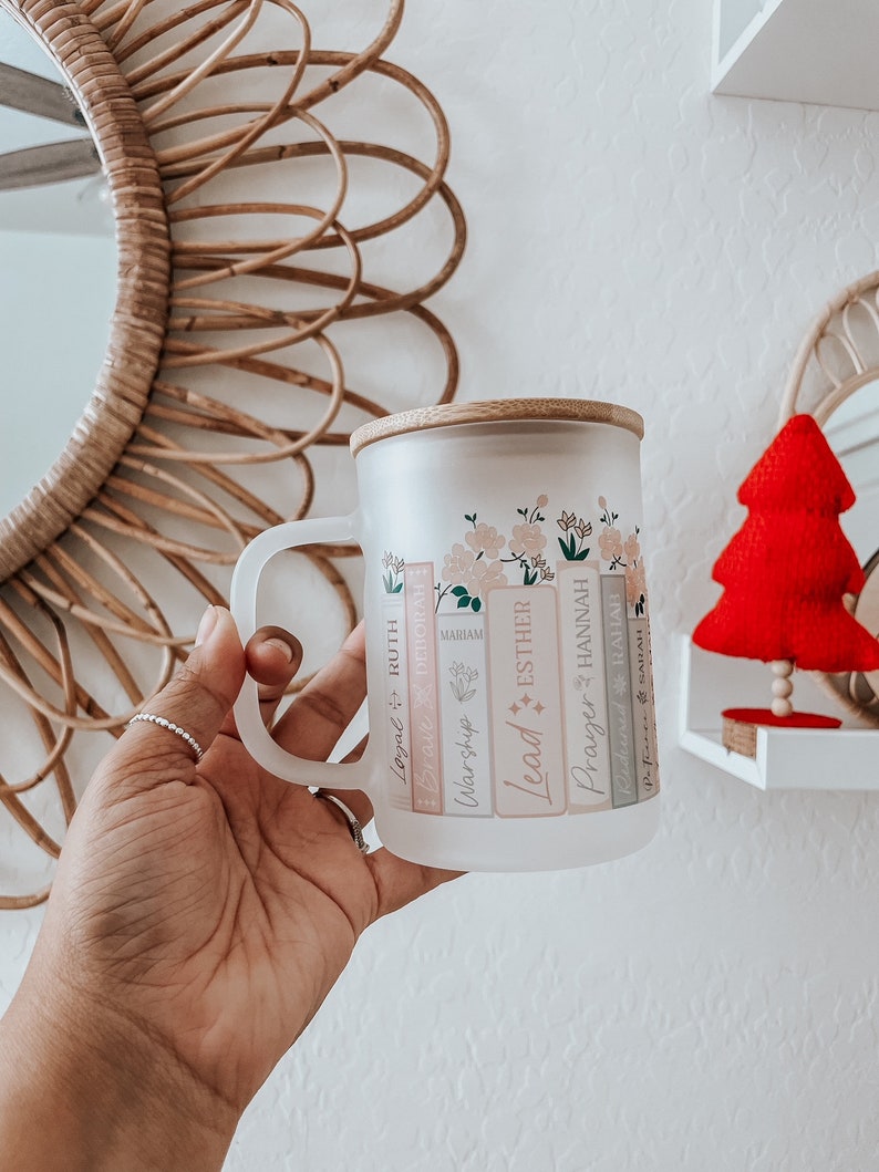 May include: A frosted glass mug with a bamboo lid and a floral design. The mug has a handle and features the words "Lead", "Worship", "Miriam", "Esther", "Prayer", "Hannah", "Legal", "Ruth", "Deborah", "Rejoice", and "Sarah" printed on the side.