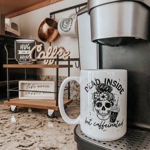 Pu&ograve; includere: Tazza da caff&egrave; in ceramica bianca con una grafica nera di uno scheletro con un chignon e fiori, e il testo "Dead Inside but caffeinated".