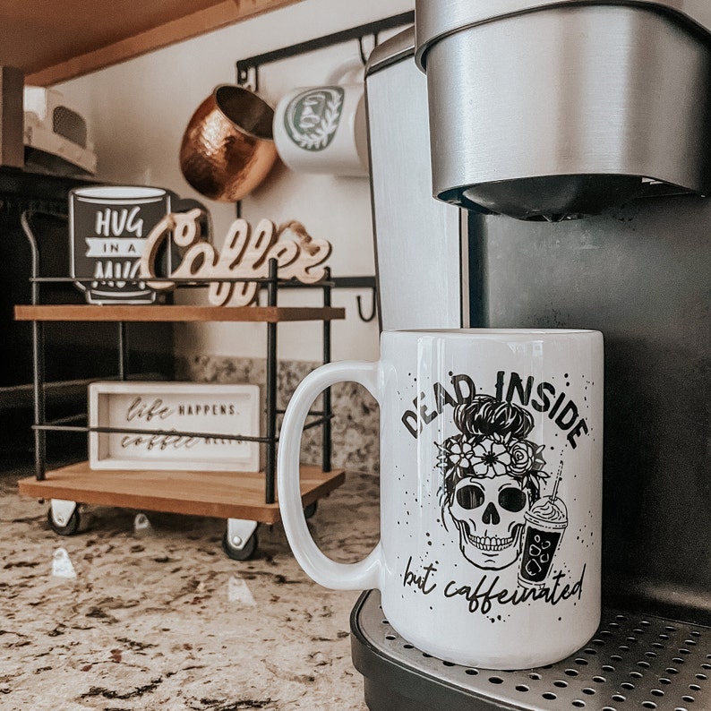 Pu&ograve; includere: Tazza da caff&egrave; in ceramica bianca con una grafica nera di uno scheletro con un chignon e fiori, e il testo "Dead Inside but caffeinated".