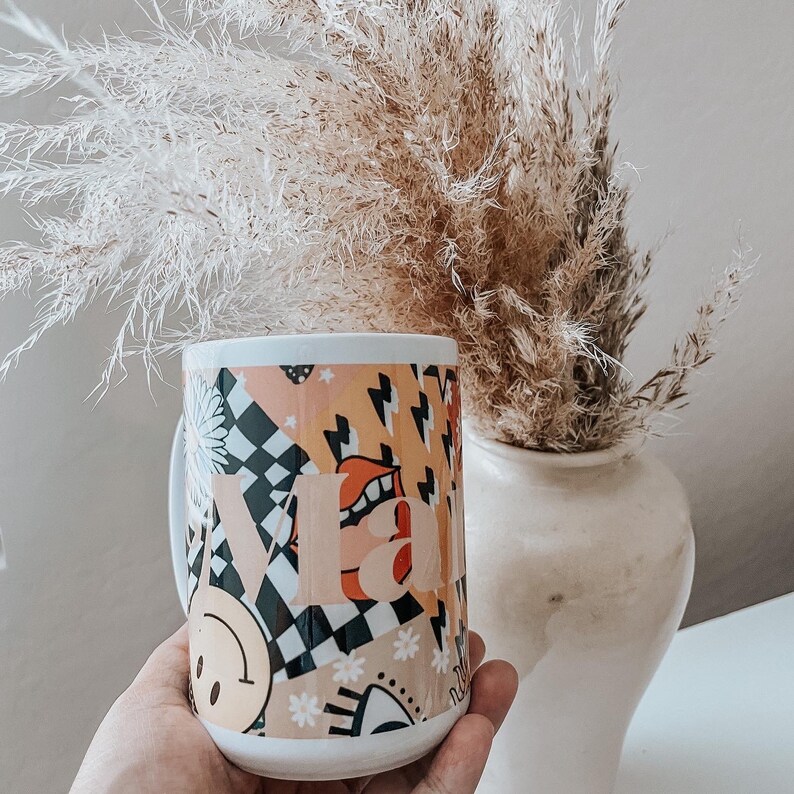 Pu&ograve; includere: Tazza in ceramica bianca con un design astratto colorato che presenta un motivo a scacchi, un viso sorridente e la parola "VIBE".