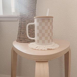 May include: A white ceramic mug with a checkered pattern in beige and white. The mug has a wooden lid and a straw. The mug is sitting on a white crocheted coaster on a light wood table.