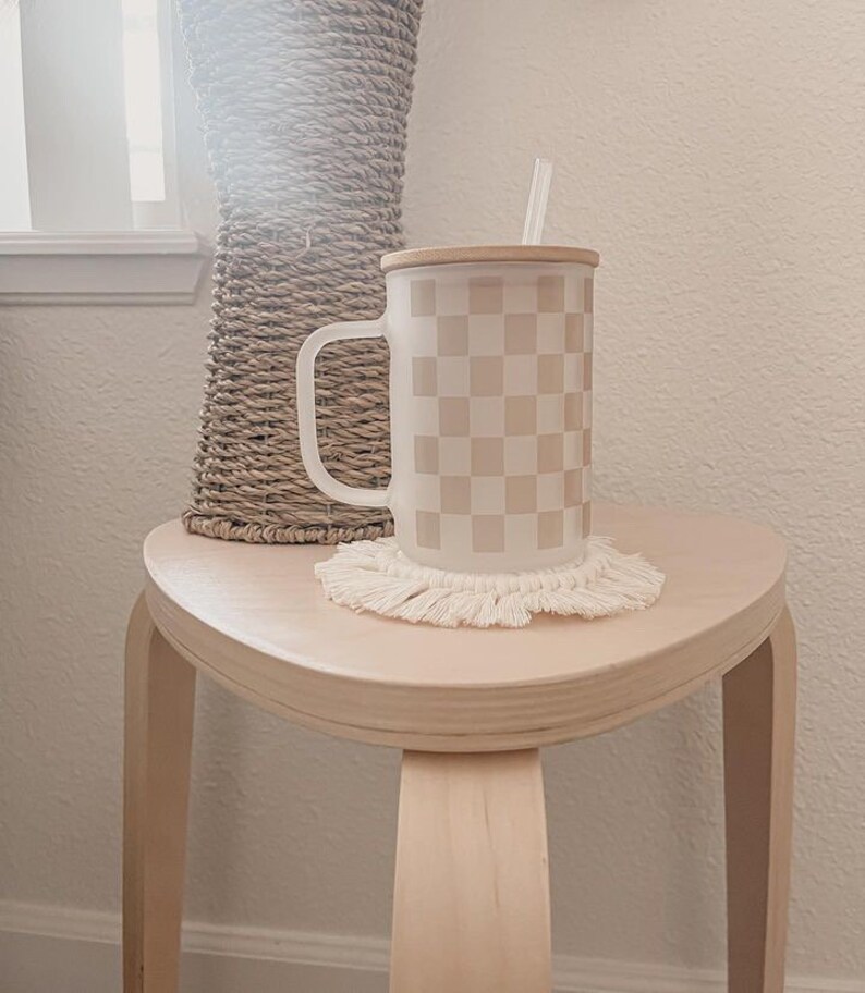 May include: A white ceramic mug with a checkered pattern in beige and white. The mug has a wooden lid and a straw. The mug is sitting on a white crocheted coaster on a light wood table.