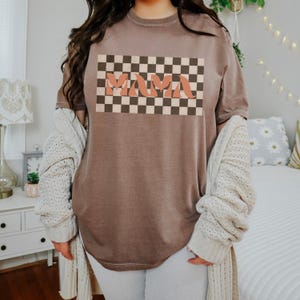 May include: A brown t-shirt with the word "MAMA" in a retro font, set against a checkered background. The shirt has a relaxed fit and is made of a soft material. The overall style is casual and vintage-inspired.