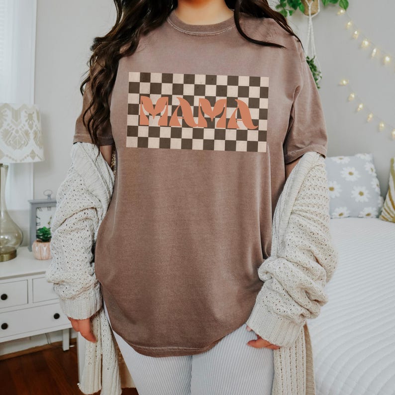 May include: A brown t-shirt with the word "MAMA" in a retro font, set against a checkered background. The shirt has a relaxed fit and is made of a soft material. The overall style is casual and vintage-inspired.