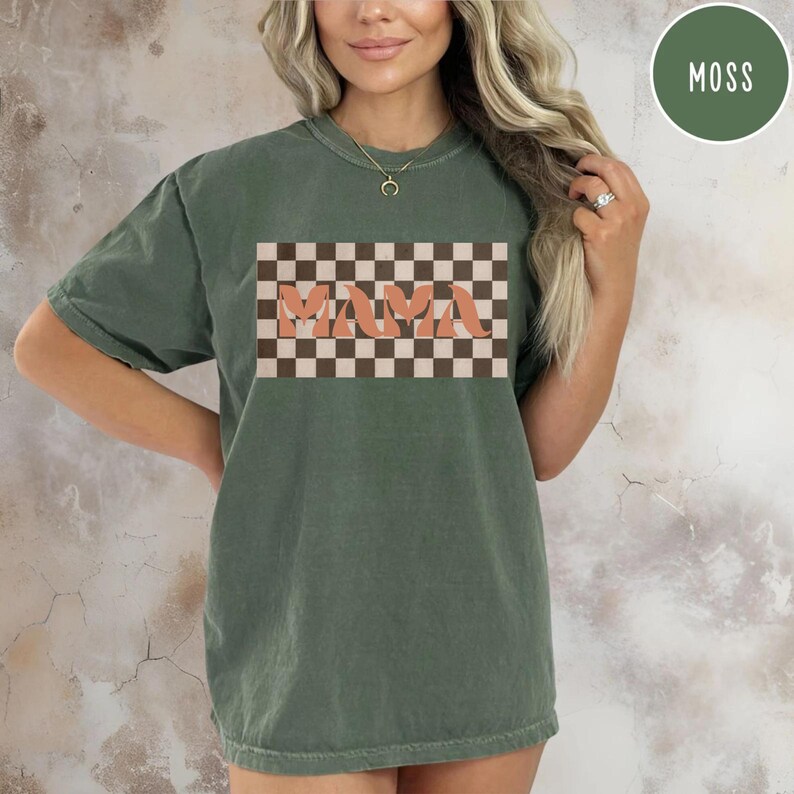 May include: Moss green t-shirt with a retro design. The shirt features the word "MAMA" in a peach-colored, retro font, set against a brown and cream checkered background. The shirt is a comfortable, casual style.