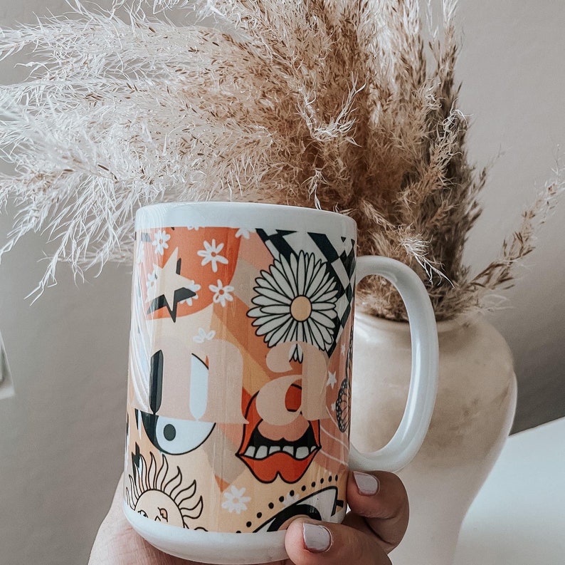 Pu&ograve; includere: Tazza in ceramica bianca con un design colorato che presenta una margherita, un sole, un occhio e la parola "mamma".