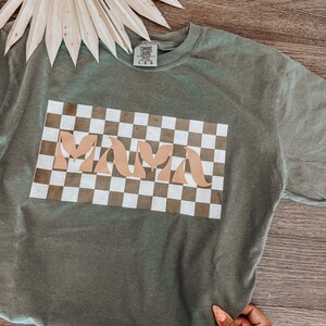 May include: Olive green crew neck t-shirt with the word "MAMA" in a tan, retro font, set against a brown and white checkered background. The shirt is displayed on a wooden surface, with a dried palm leaf in the background.