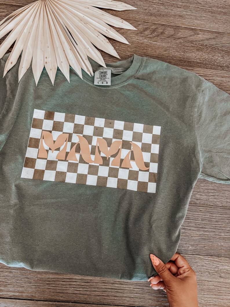 May include: Olive green crew neck t-shirt with the word "MAMA" in a tan, retro font, set against a brown and white checkered background. The shirt is displayed on a wooden surface, with a dried palm leaf in the background.