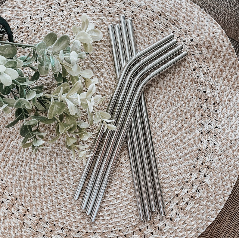 May include: A set of silver reusable drinking straws with a bend at the top. The straws are arranged on a woven straw mat.