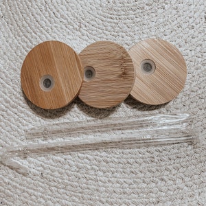 May include: Three round, wooden lids with a small hole in the center. The lids are made of light brown bamboo and are arranged in a row. Two clear glass straws are lying on a woven surface.
