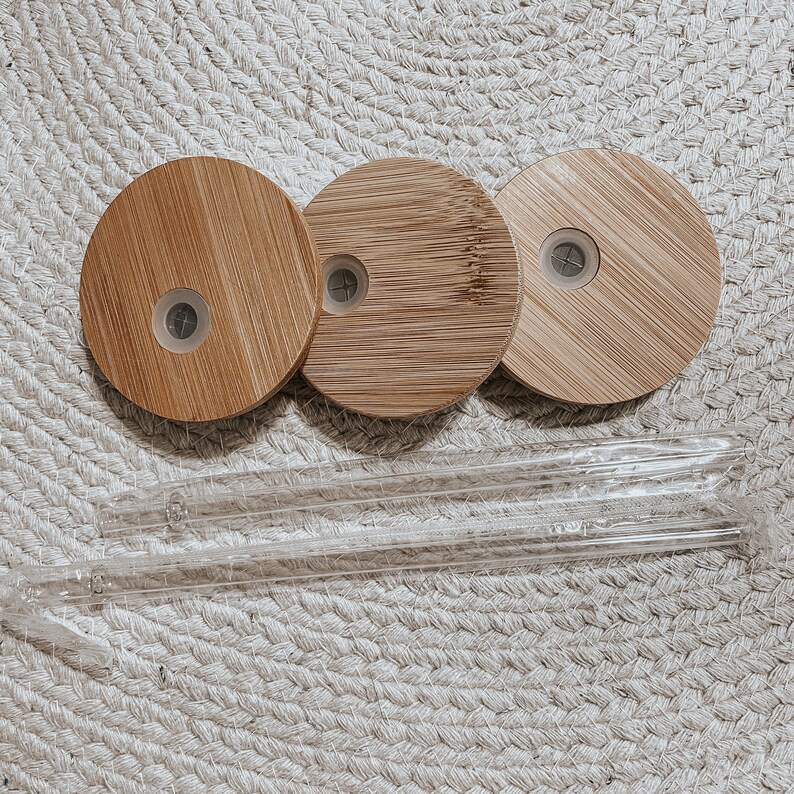 May include: Three round, wooden lids with a small hole in the center. The lids are made of light brown bamboo and are arranged in a row. Two clear glass straws are lying on a woven surface.