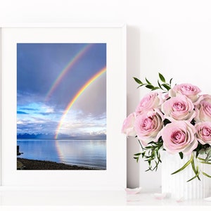 Puede incluir: Una fotografía enmarcada de un doble arcoíris sobre una tranquila masa de agua. El arcoíris se extiende sobre un cielo nublado, reflejándose en el agua. Un ramo de rosas rosas en un jarrón blanco se encuentra a la derecha.