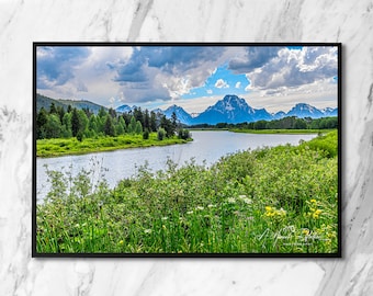 Puede incluir: Fotografía de paisaje enmarcada que muestra un río tranquilo que serpentea a través de un prado verde. Montañas con picos nevados se alzan a lo lejos bajo un cielo azul nublado. La escena está enmarcada en un borde negro.