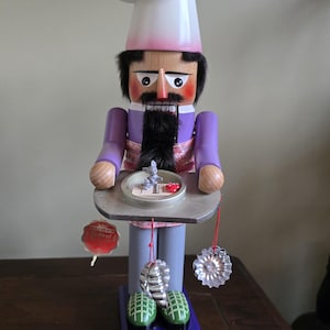 May include: A wooden nutcracker dressed as a chef, wearing a pink and white hat, a purple jacket, and a grey apron. He is holding a tray with a small red and white cake on it. There are three silver cookie cutters hanging from the tray.