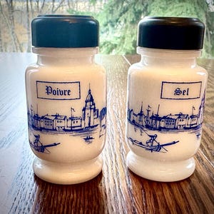 May include: Two white ceramic salt and pepper shakers with black lids. The shakers have blue and white illustrations of a city skyline and a boat. The shakers are labelled "Sel" and "Poivre".