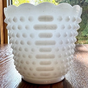 May include: A white hobnail milk glass vase with a scalloped rim. The vase has a textured surface with raised dots and horizontal rectangular indentations. The vase is sitting on a wooden surface.