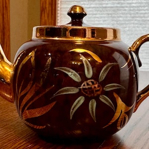 May include: A brown ceramic teapot with gold detailing on the spout, handle, lid, and rim. The teapot has a floral pattern with gold and white accents. This teapot is likely for serving tea.
