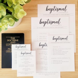 May include: A collection of white cards with the words "baptismal covenants" in elegant script, alongside a black book titled "The Doctrine and Covenants." The cards list promises and covenants, with a small green plant in the background.