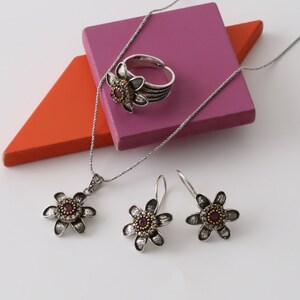 Puede incluir: Un conjunto de collar, anillo y pendientes de plata en forma de flor con una piedra roja en el centro de cada pieza. Las joyas se muestran sobre un fondo rosa y naranja.