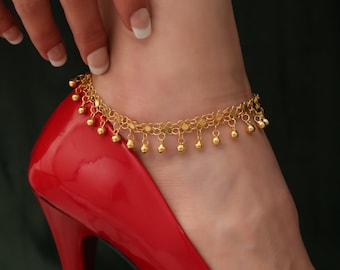 May include: A gold ankle bracelet with small round charms. The bracelet is on a woman's ankle, and she is wearing a red high-heeled shoe.