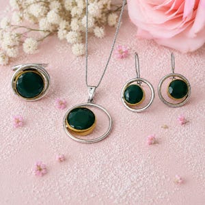May include: A jewelry set consisting of a necklace, a ring, and a pair of earrings. Each piece features a round, faceted dark green stone set in a gold-tone bezel, framed by a larger, thin silver-tone circular metal ring design.