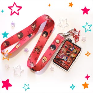Peut inclure: Un cordon rouge orné d'un motif répétitif de visages de personnages de dessin animé et d'étoiles, attaché à une breloque rectangulaire en acrylique. La breloque présente une grille d'illustrations rappelant un écran de smartphone avec un fermoir en métal.
