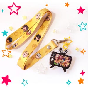 May include: A yellow lanyard featuring a repeating pattern of various illustrated character faces and circular motifs. Attached to the lanyard is a silver-toned metal clasp holding a black acrylic charm shaped like a vintage television set.