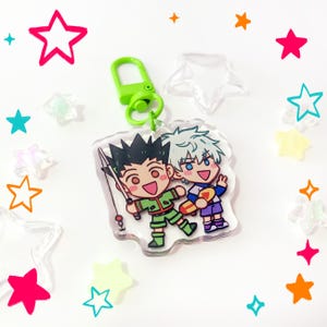 May include: A clear acrylic keychain featuring a cartoon illustration of two characters, one with spiky black hair holding a fishing rod and one with white hair holding a skateboard, attached to a bright lime green plastic lobster clasp.