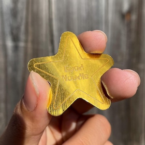 May include: A gold-colored star-shaped token with a brushed metallic finish. The center of the star features the engraved text "Good Noodle" in a simple sans-serif font, surrounded by a thin engraved outline that follows the star's shape.