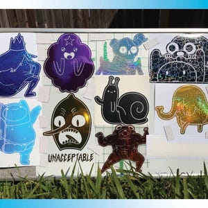 Peut inclure: Une collection d'autocollants color&eacute;s repr&eacute;sentant des personnages de la s&eacute;rie anim&eacute;e Adventure Time. Les autocollants sont affich&eacute;s sur une surface blanche et comprennent divers motifs, tels que le Roi des Glaces et Finn.
