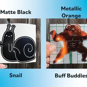 Peut inclure: Deux mod&egrave;les d'autocollants sont pr&eacute;sent&eacute;s. L'un est un escargot noir mat avec le mot "Snail" en dessous. L'autre est une grenouille muscl&eacute;e orange m&eacute;tallique avec les mots "Buff Buddies" en dessous. Le fond est une surface en bois.