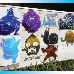 Peut inclure: Une collection d'autocollants holographiques repr&eacute;sentant des personnages de dessins anim&eacute;s. Les autocollants sont de diff&eacute;rentes couleurs, notamment le bleu, le violet, l'or et le noir. Le mot "UNACCEPTABLE" est imprim&eacute; sur l'un des autocollants.