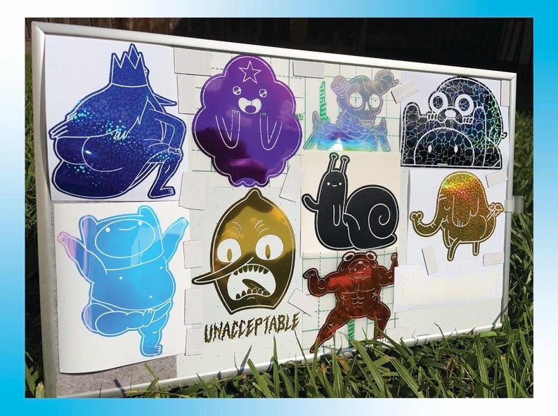 Peut inclure: Une collection d'autocollants holographiques repr&eacute;sentant des personnages de dessins anim&eacute;s. Les autocollants sont de diff&eacute;rentes couleurs, notamment le bleu, le violet, l'or et le noir. Le mot "UNACCEPTABLE" est imprim&eacute; sur l'un des autocollants.