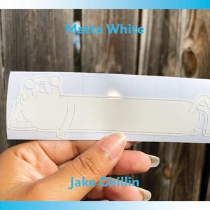 Peut inclure: Autocollant en vinyle blanc mat repr&eacute;sentant un personnage de dessin anim&eacute; dans une pose d&eacute;tendue. L'autocollant porte le texte "Jake Chillin" et "Matte White". L'autocollant est tenu par une main.