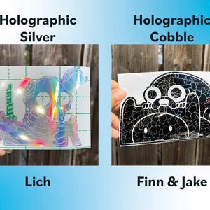 Peut inclure: Deux autocollants holographiques sont pr&eacute;sent&eacute;s. L'un est un design Lich argent&eacute;, et l'autre est un design Finn & Jake avec un effet pav&eacute;. Les autocollants sont pr&eacute;sent&eacute;s sur un fond de cl&ocirc;ture en bois.
