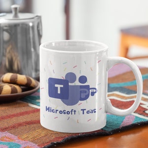 Puede incluir: Taza de cerámica blanca con el logotipo de Microsoft Teams y las palabras "Microsoft Teas" en azul. La taza tiene un asa y está decorada con confeti de colores. Un plato de galletas y una tetera de metal están en el fondo.