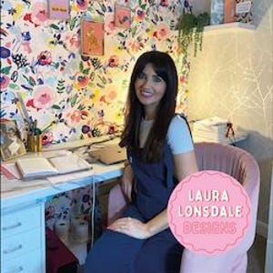 May include: A woman sits at a desk in a room with floral wallpaper. The desk has a laptop, open book, and stationery. A pink chair and a pink logo with the text "LAURA LONSDALE DESIGNS" are also visible.