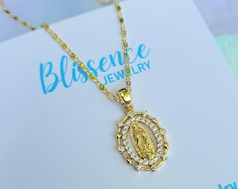 May include: Gold chain necklace with a pendant featuring a depiction of the Virgin Mary surrounded by clear cubic zirconia stones. The pendant is oval shaped and has a detailed design. The necklace is displayed on a white card with the text "Blissence Jewelry" and social media icons.