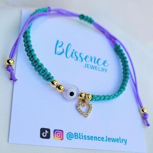 May include: A green and purple string bracelet with a gold evil eye charm and a gold heart charm. The bracelet is on a white card with the text "Blissence Jewelry" and social media handles.