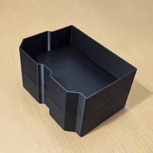 May include: A matte black rectangular storage tray with a stepped, notched side profile. The container features a deep, open interior with clean, sharp edges and a solid, uniform construction, designed for organizing small items on a flat surface.