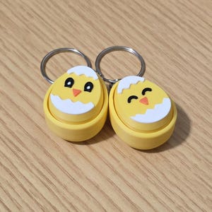 May include: Two yellow chick-shaped keychains with silver key rings. Each keychain features a chick emerging from a cracked eggshell. The chicks have black eyes and orange beaks, with white accents on the eggshell.